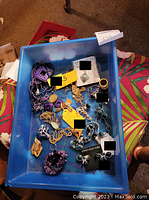 Photo of a blue container holding various items of costume jewelry in gold, silver tones, and purple, including necklaces, rings, earrings, bracelets, some with purple and turquoise stones.