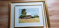 Front view of watercolor painting showing barn, trees, grass, and sky in a gold frame with white mat.