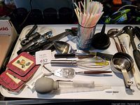 Photo of various kitchen utensils, measuring tools, and coasters arranged on a white surface, including ladles, measuring cups, a manual citrus juicer, coasters, and a glass with straws.