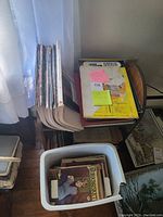 Bucket with a number of vintage 1970s Workbasket magazines with illustrated covers placed on a wooden floor near the bookcase.
