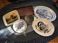 Five assorted serving trays displayed on a fabric surface, including a winter scene platter, Walter turkey platter, floral tray, vintage style framed tray, and wooden tray