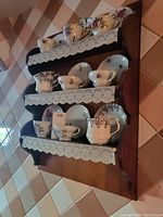 Three-tier wooden shelf with lace-trimmed shelves holding various tea cups and saucers with floral patterns and gold trim.