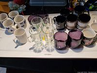 Multiple ceramic mugs with floral and solid blue or pink designs, clear glass mugs and glasses, and two dark purple tinted glass candle holders on a table surface.