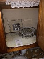 Photo showing stacked set of 4 vintage Currier and Ives winter plates with scalloped edges and gold trim holding a lot number tag inside a wooden cabinet.