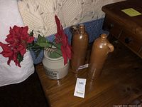 Photo showing two vintage stoneware jugs side by side on a wooden surface with some flowers in a ceramic container nearby.