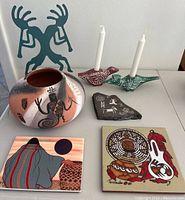 Entire lot showing pottery pot, two candleholders each holding white candle, two signed art tiles, and petroglyph slab on a white table.