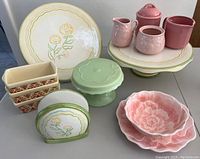 Image showing collection of ceramic serving pieces including cake plates with floral motifs, mini loaf dishes, cake pedestal, pink flower-shaped plates, and cream and sugar set.