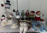 Wide photo showing all assembled Christmas decor items on a table including metal snowman frame, plush snowmen, nutcrackers, plush pudding, crocheted angel, wreath with plush bear, and teddy bears.