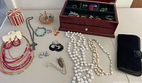 Overview of various costume jewelry including necklaces, bracelets, earrings, some faux pearl strands and silver tone pieces. Includes a red and white tin container and a wooden jewelry box.