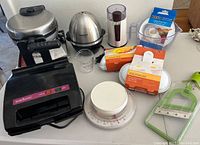 Photo of assorted kitchen appliances and tools including Cuisinart Egg Central egg cooker, waffle makers, coffee grinder, kitchen scale, and other items on a table.