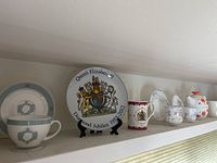 Shelf displaying multiple British Royalty collectible plates and English porcelain mugs and cups, including a Queen Elizabeth II Diamond Jubilee commemorative plate.