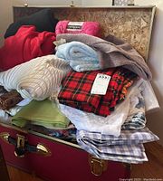 Image showing assorted fabrics and fleeces stacked inside a red trunk. Variety of materials and colors visible including pink fleece, plaid, gingham, textured white, and green fabrics.