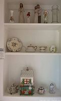 Overview of three shelves showing Willow Tree style figurines on upper shelf, porcelain plate and teapots with floral designs on middle shelf, and various trinket boxes including ceramic houses and small floral teapots on lower shelf.
