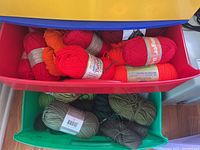 Plastic storage drawers containing mostly full yarn skeins in reds and oranges in the red drawer, greens in the green drawer, and white, purple, and green in the yellow drawer.