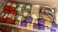 Bin with skeins of orange, green variegated, cream, and purple yarn, also pattern booklet visible