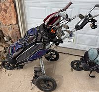 Golf bag with clubs and Bag Boy rolling cart shown from side angle on driveway in front of garage door.