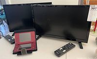 Two 18-inch Insignia TVs with black remote controls and a red Nintendo DS XL handheld gaming console in a black protective case displayed on a table.