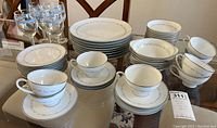 Photo showing the full 44-piece Noritake Brooklane porcelain dishware set on a glass table including plates, cups, saucers, bowls, and blue-rimmed glasses.