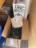Original Thigh Master exercise device in its original box and green bungee cords inside a cardboard box with a beige canvas bag