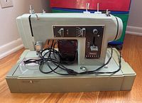 Sears Kenmore sewing machine sitting on the sewing machine base with power cord and foot pedal attached. Machine is light green with control dials on the front panel.