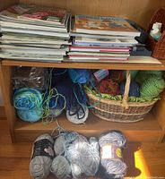Books, magazines stacked on top shelf above yarn skeins. Yarn skeins in basket and loose on bottom shelf in various colors and sizes.