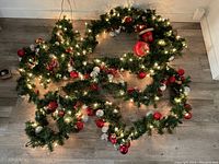 Full view of the green Christmas garland decorated with red and silver ornaments and lit with miniature string lights arranged on the floor.