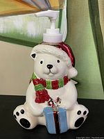 Front view of ceramic polar bear soap dispenser holding a blue gift with a red ribbon, wearing red Santa hat and green scarf.