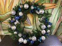 Full view of the circular green artificial pine Christmas wreath decorated with blue, white, silver, and metallic blue baubles and different shaped ornaments, showing overall condition and presentation.