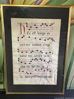 Full front view of framed manuscript showing gold-tone frame, black matte, vellum sheet with red initial and neumes