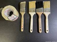 Four paint brushes and two rolls of masking tape on black surface.