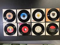 Photo showing 8 vintage 45 RPM vinyl records arranged in a 4x2 grid on a dark background. All records have visible labels and some have paper sleeves.
