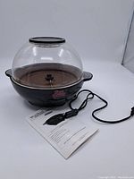Front view of the West Bend Stir Crazy popcorn popper showing black base, clear dome lid, power cord, and instruction manual.