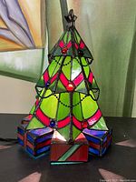 Front view of stained glass Christmas tree lamp showing green, pink, and red glass panels with hanging chain garlands and glass bead ornaments. Presents at the base visible.