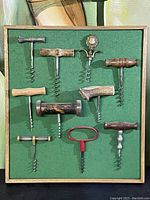 Full view of 10 assorted vintage corkscrews arranged on green felt inside a wood frame.
