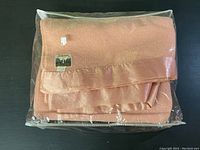 Full view of the folded pink wool blanket wrapped in plastic, showing satin trim and size label.