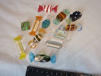 Twelve decorative blown glass candy pieces arranged on a white background with a black measuring ruler showing size scale.