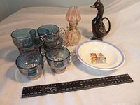 Photo showing the mini oil lamp, duck jug, six blue-tinted glass coffee cups, and child's bowl with character illustrations beside a ruler for size reference.