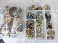 Clear plastic container with multiple compartments holding various costume jewelry pieces including rhinestone brooches, blue evil eye beads, metal rings, and faux pearl strands.