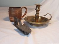 Photo of three assorted metal items: a brass finger candle holder, a copper cup, and a cast iron hand shovel door knocker on a white surface.