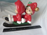 Side view of ceramic figure showing two children on sled with detailed red outfits and faces, ruler for scale.