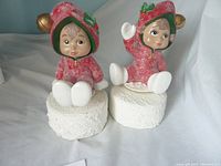 Front view of two Christmas musical figurines with childlike faces, dressed in red textured winter outfits, sitting on white snow-like bases, one with a raised hand holding a jingle bell.