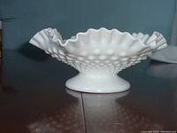 Side view of white milk glass Fenton hobnail bowl with crimped ruffled edge and footed base on a wooden surface.