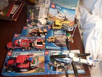 Photo showing multiple assembled and partially assembled Lego City vehicles including tractor, truck with crane platform, helicopter, and instruction booklets.