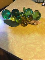 Top and angled side view of multi-size, multi-color lucite grapes arranged on tabletop showing translucent spheres in green, dark green, and amber colors with attached artificial leaves.