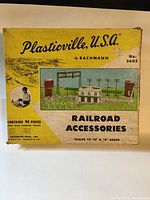 Original yellow box with Plasticville U.S.A. Railroad Accessories label, showing set No. 5605 and artwork with train station and landscape. Box shows wear.