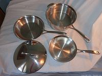 Three stainless steel pans of varying sizes with long handles, placed on a fabric surface, alongside a matching heavy lid.