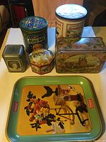 Photo showing all tins arranged on a surface with the large rectangular Coca-Cola tray prominently in front, and five assorted collectible tins including Milky Way and Canadian history tin.