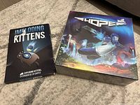 Photo showing Imploding Kittens expansion box and Hope board game box side by side on carpet.