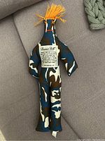 Front view of the Dammit Doll stress reliever showing full doll with camo print fabric, orange yarn hair and label on torso.