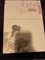 Top of the decorative beige box featuring a detailed painting of an Asian woman holding a fan, with East Asian characters and brand name Alessir.